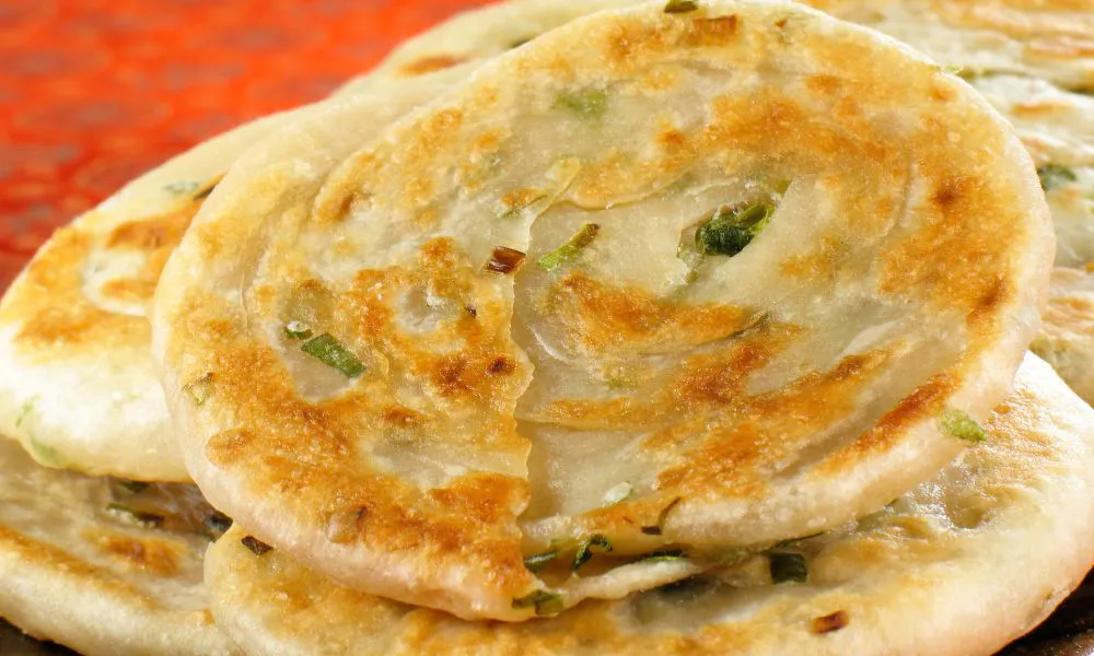 Stacked golden pan-fried scallion pancakes with layered flaky dough and fresh green scallion bits on red surface at Princess Dumpling, a Asian Restaurant in Rowland Heights